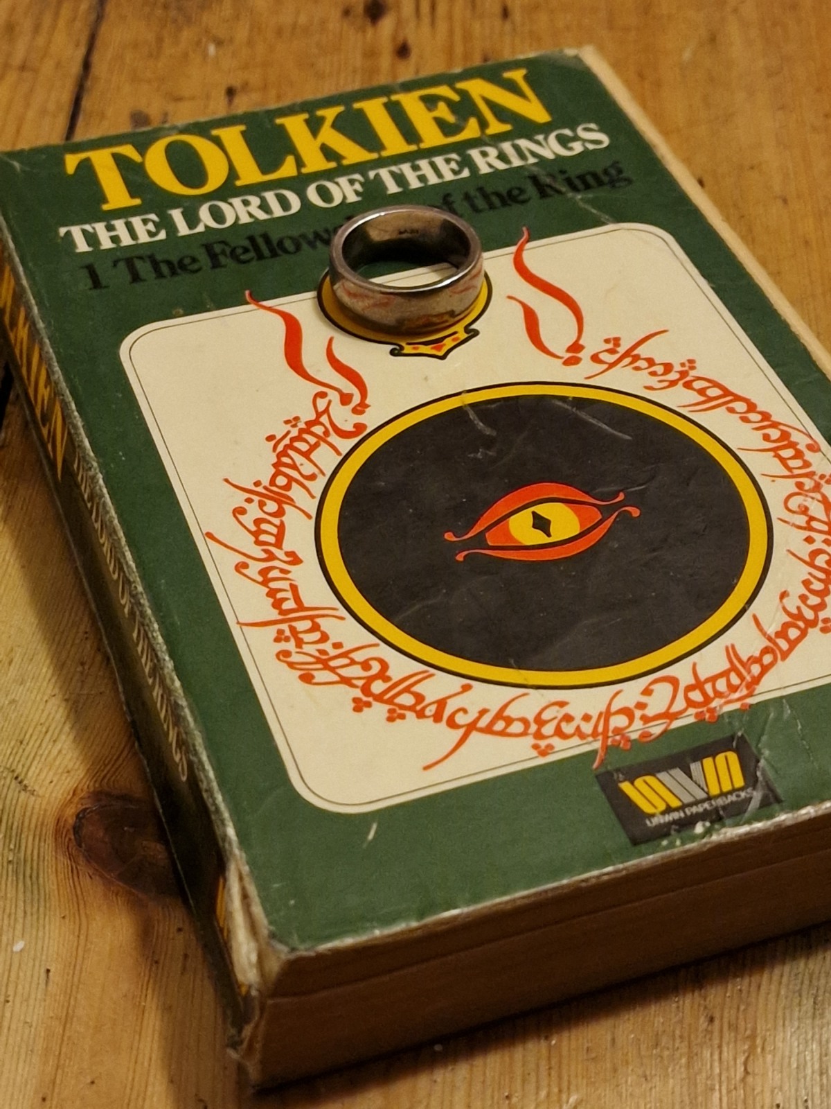 An old copy of Lord of the Rings sits on the floor, with a ring on top. The cover is green with a big black circle with a stylised eye in the centre, around this is red fancy script