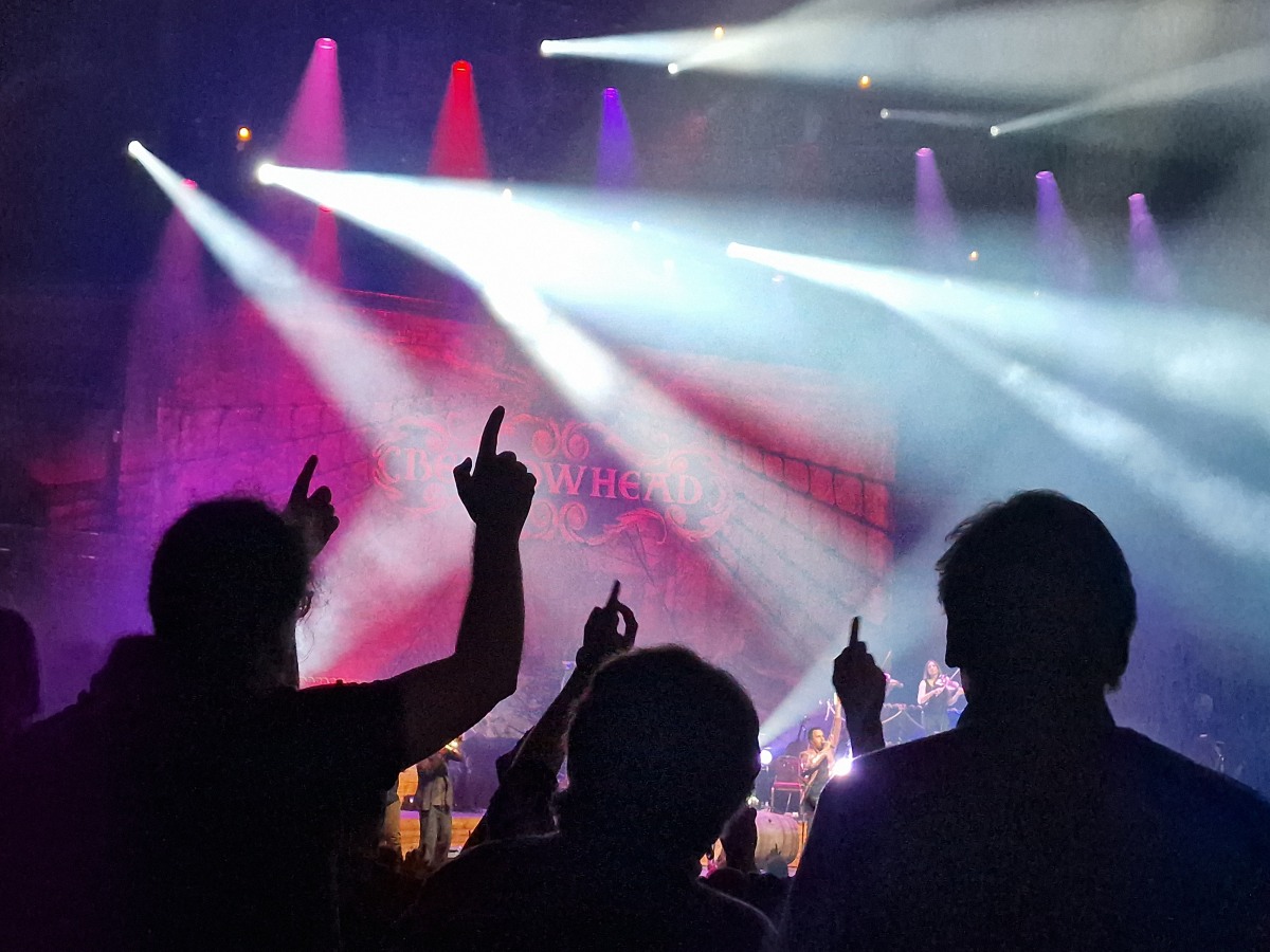 Three people are silhouetted against bright spot lights, they are dancing and two have their hands pointing up towards the ceiling.