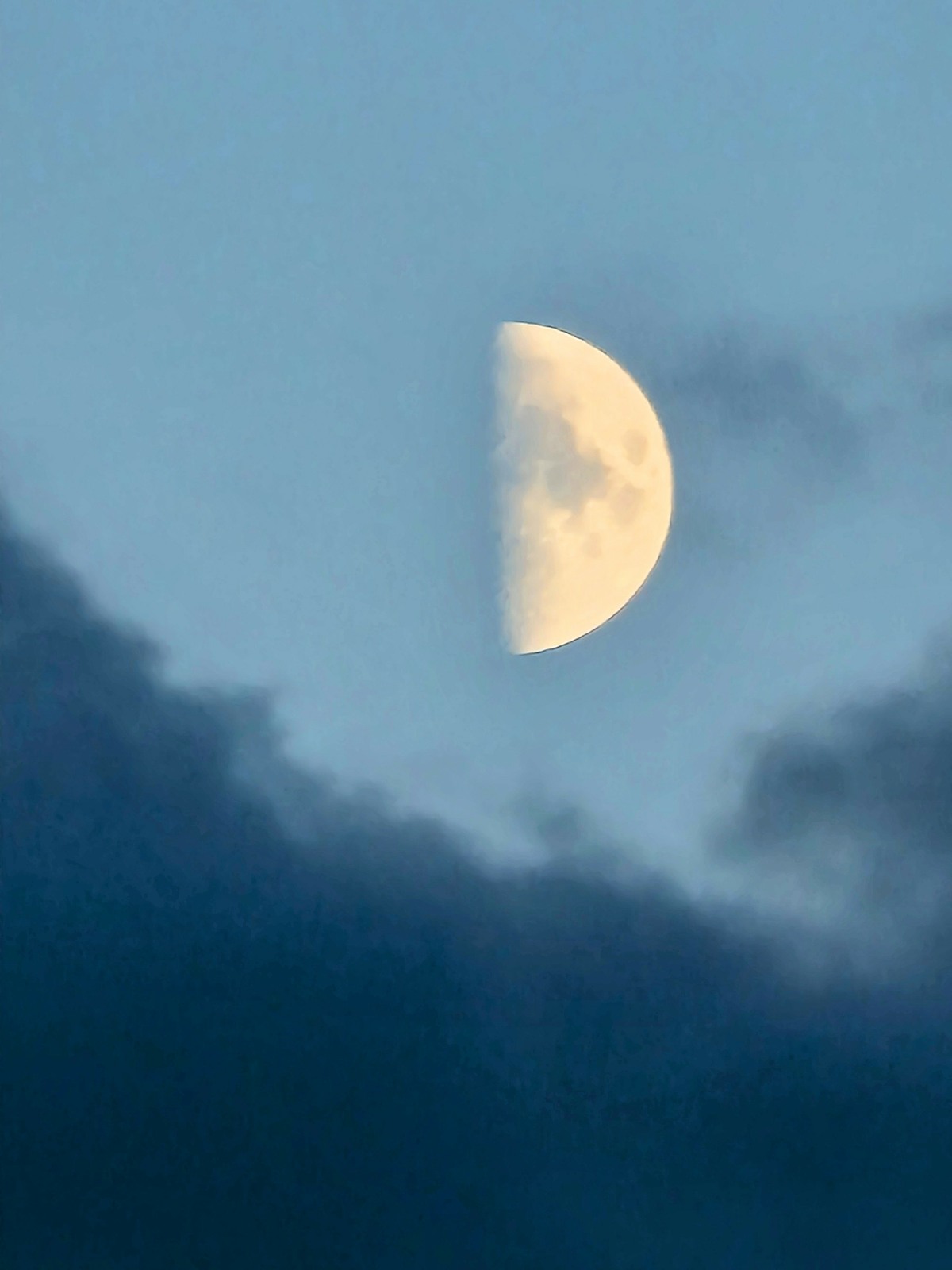 The moon, halfway between new moon and full moon, in a blue sky, with black clouds across the bottom of the picture.