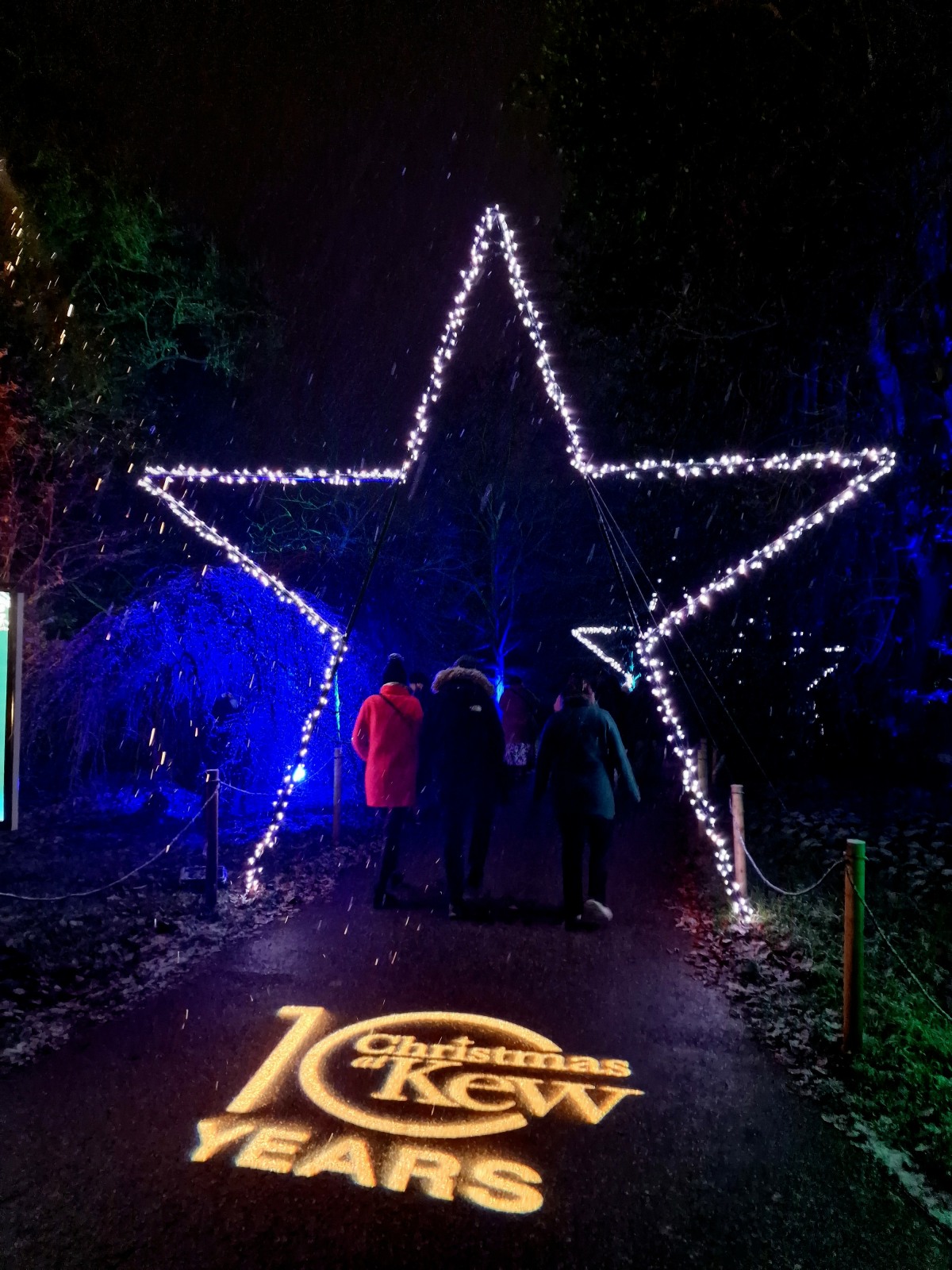 An outline of a Star straddles a walkway, three people walking underneath.  One is wearing a red coat. On the ground is written 10 years, Christmas at Kew.