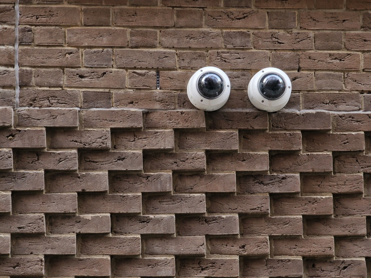 A brick wall, with patterned bricks, has two  security camera domes that look like a pair of googly eyes.