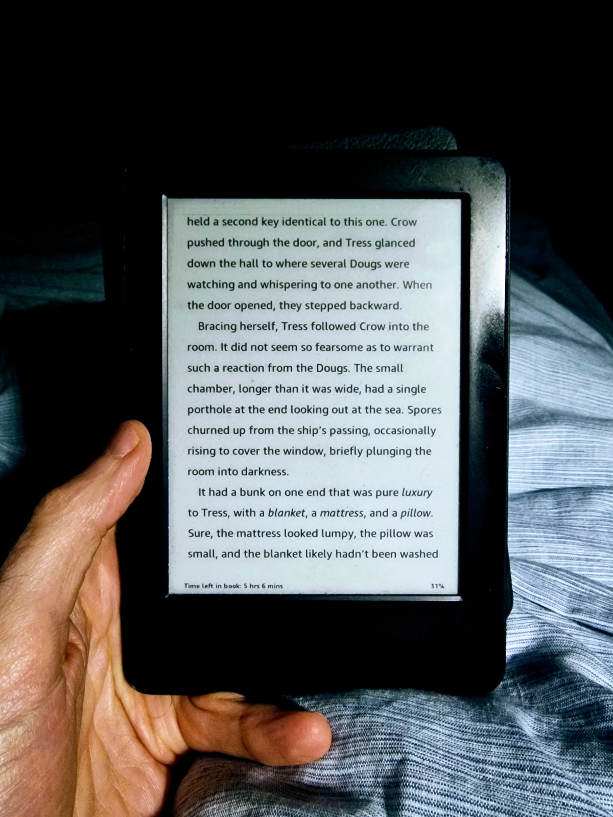 A hand holds a kindle against a dark background and a blue duvet.