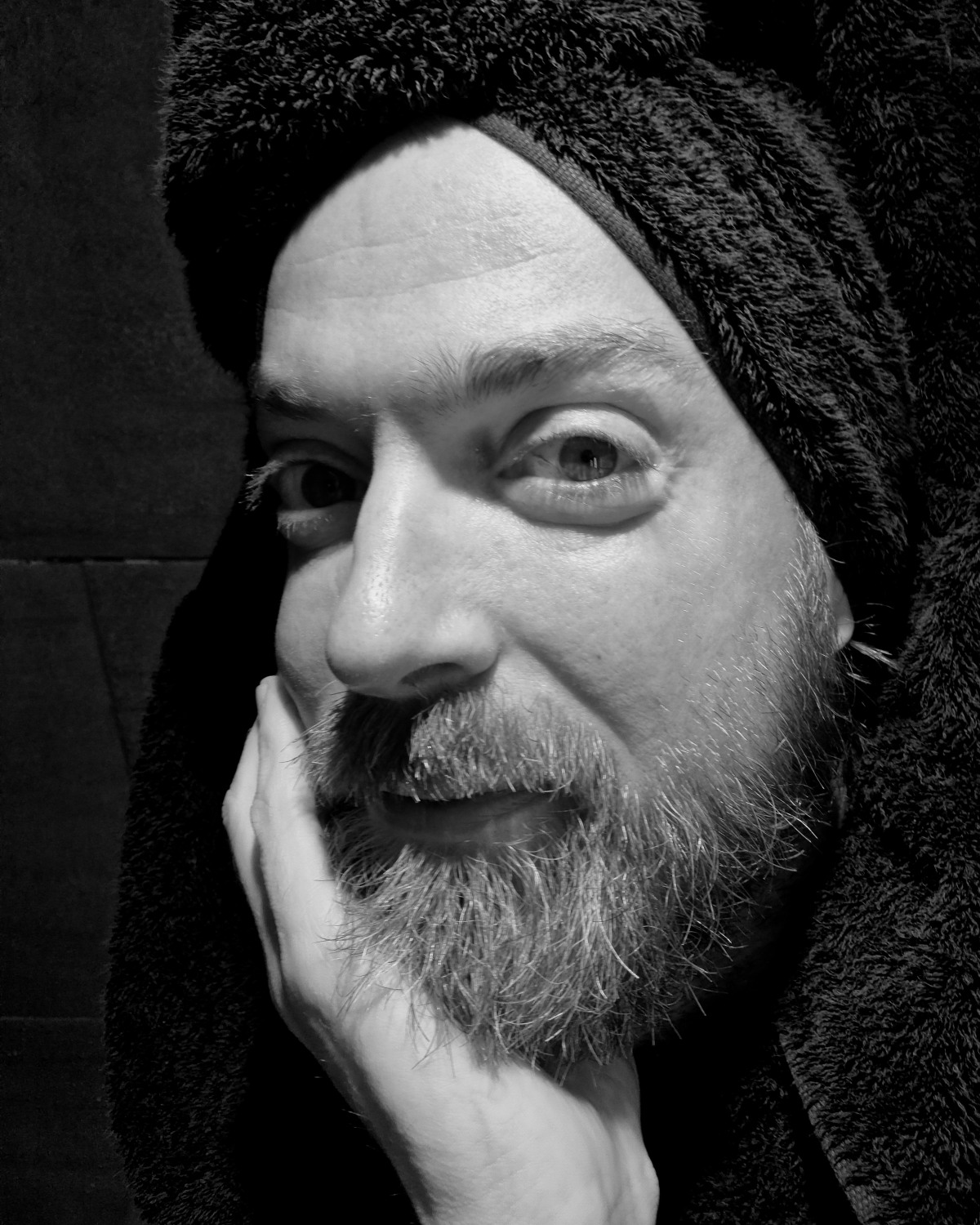 Black and white portrait, man with beard, head wrapped in a dark towel, rests his chin in his hand and looks directly out of the picture, a slight smile.